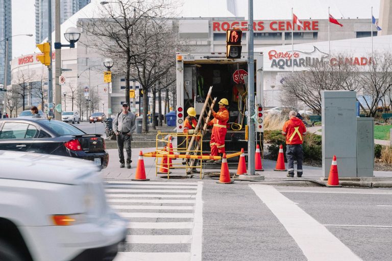 Full body of workers in red uniforms and yellow helmets carrying wooden ladder in street near automobile road